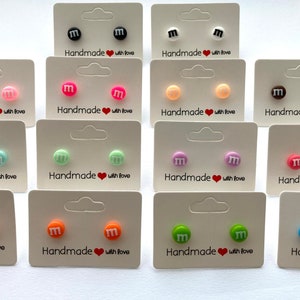May include: Assortment of colorful stud earrings featuring a white "M" on a round background. The earrings are displayed on white cards with the text "Handmade with love".