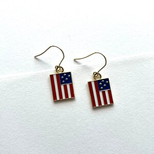 May include: A pair of gold earrings with a red, white, and blue American flag design. The flags have seven stars.