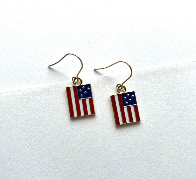 May include: A pair of gold earrings with a red, white, and blue American flag design. The flags have seven stars.