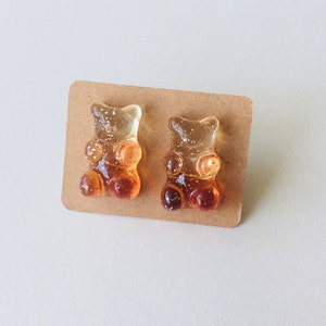 May include: A pair of gummy bear earrings, one is orange and the other is yellow. The earrings are on a brown card.
