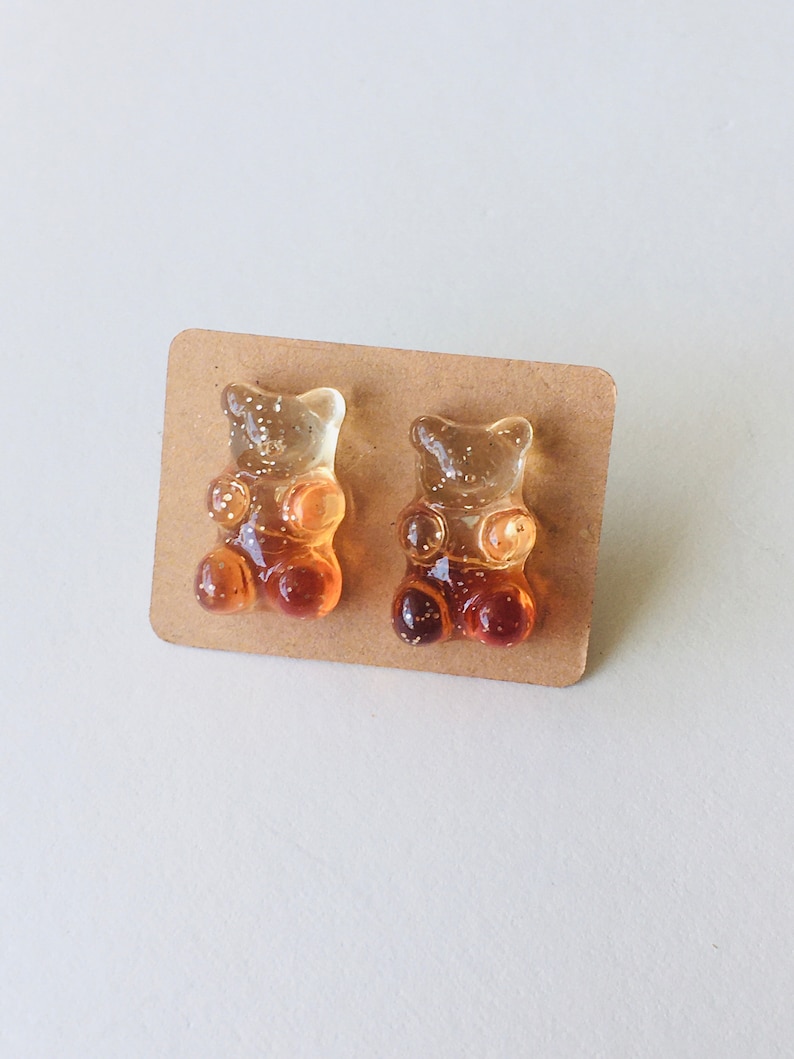 May include: A pair of gummy bear earrings, one is orange and the other is yellow. The earrings are on a brown card.