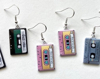 May include: Four pairs of cassette tape earrings. Two pairs are green with the text "Good Vibes", one pair is pink with the text "Best of the 80's", and one pair is gray with the text "Just Groove".
