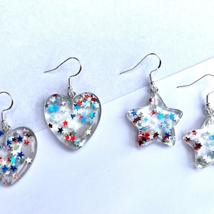 May include: Clear resin earrings in heart and star shapes, filled with red, white, and blue star-shaped glitter. The earrings have silver-colored fish hook ear wires. The earrings are a patriotic theme.