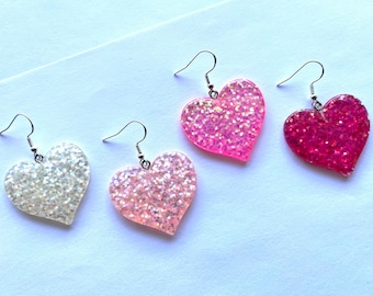May include: Three pairs of heart-shaped earrings with glitter. The earrings are in shades of pink, white, and red.