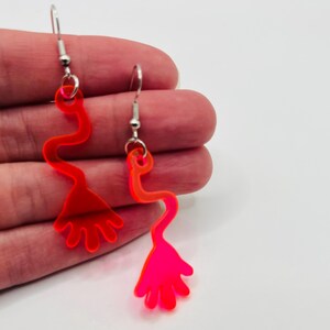 May include: A pair of neon pink acrylic earrings shaped like a hand with a long, thin arm. The earrings are on a silver hook.