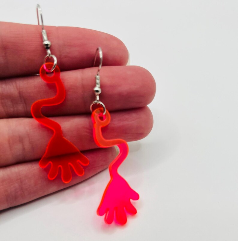 May include: A pair of neon pink acrylic earrings shaped like a hand with a long, thin arm. The earrings are on a silver hook.