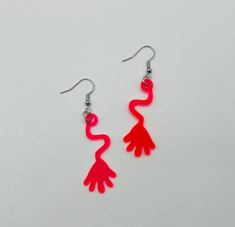May include: A pair of pink acrylic earrings shaped like a hand with a wavy line leading up to it.