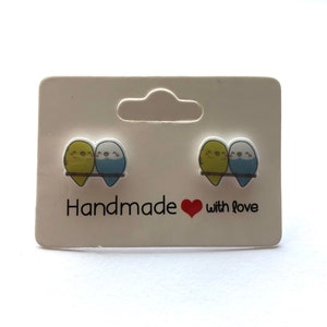 May include: A pair of handmade earrings featuring two small blue and yellow birds on a branch. The earrings are on a white card with the text "Handmade with love".