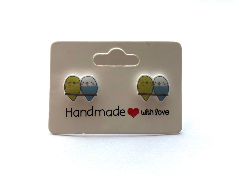 May include: A pair of handmade earrings featuring two small blue and yellow birds on a branch. The earrings are on a white card with the text "Handmade with love".