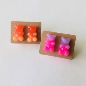 May include: Two pairs of gummy bear earrings. One pair is orange and yellow, the other is pink and purple.