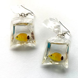 May include: A pair of earrings featuring a yellow fish inside a clear plastic bag charm. The earrings are silver and have a hook closure.