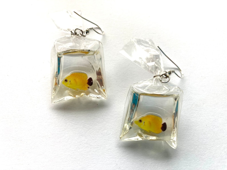May include: A pair of earrings featuring a yellow fish inside a clear plastic bag charm. The earrings are silver and have a hook closure.