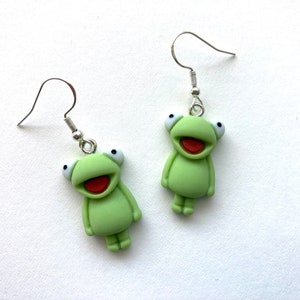 May include: A pair of green frog dangle earrings with silver hooks. The frogs have white eyes and red mouths.