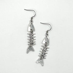May include: A pair of silver fishbone earrings with a dangling design. The earrings are made of metal and have a silver finish.