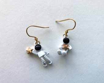 May include: A pair of gold astronaut earrings with a white astronaut holding a gold star and a white astronaut holding a gold star. The astronauts have black eyes.