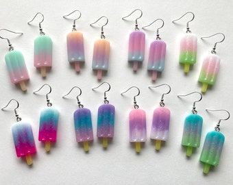 May include: A set of 12 pairs of popsicle dangle earrings. The earrings are made of resin and feature a variety of colors, including pink, blue, green, and purple. Each popsicle has a glitter finish.