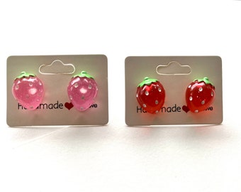 May include: Two pairs of strawberry stud earrings on white cards. One pair is pink and the other is red.