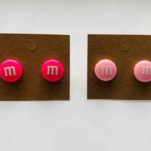 K&ouml;nnte beinhalten: Zwei Paar Ohrringe, ein Paar ist rosa und das andere ist pink. Beide Paare haben ein wei&szlig;es "M" auf der Vorderseite.