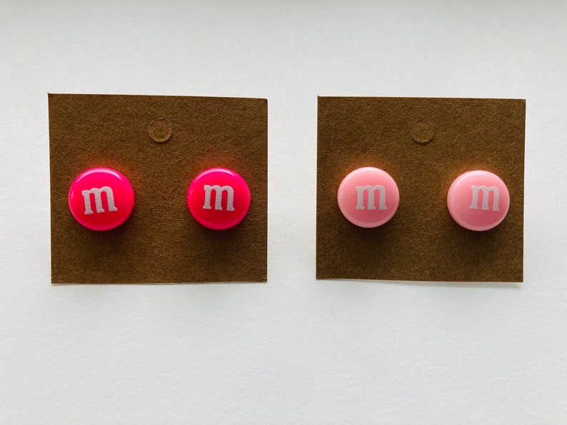 K&ouml;nnte beinhalten: Zwei Paar Ohrringe, ein Paar ist rosa und das andere ist pink. Beide Paare haben ein wei&szlig;es "M" auf der Vorderseite.