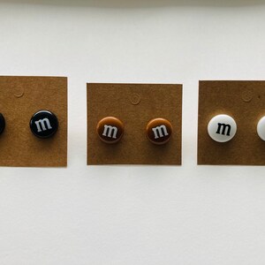 K&ouml;nnte beinhalten: Drei Paar Ohrringe mit braunem, schwarzem und wei&szlig;em Hintergrund. Jedes Paar hat einen runden Ohrstecker mit dem Buchstaben "M" in Wei&szlig;.