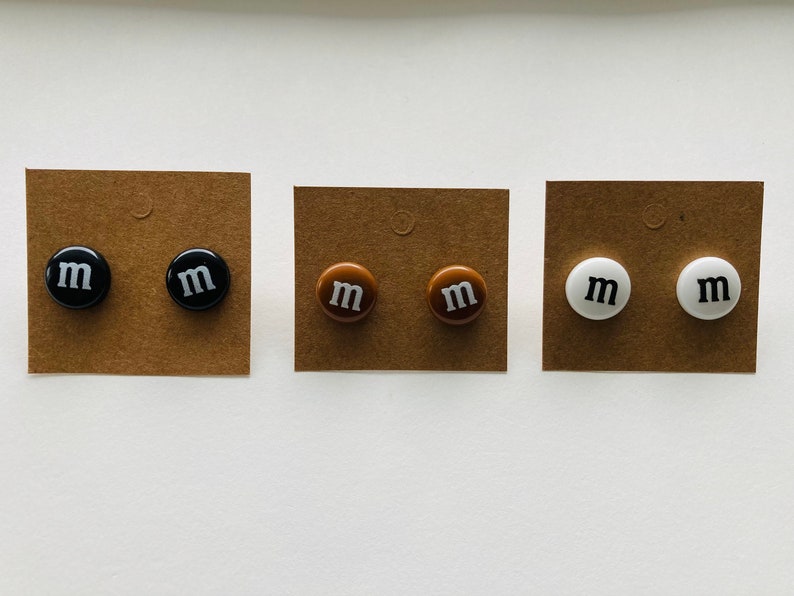 K&ouml;nnte beinhalten: Drei Paar Ohrringe mit braunem, schwarzem und wei&szlig;em Hintergrund. Jedes Paar hat einen runden Ohrstecker mit dem Buchstaben "M" in Wei&szlig;.