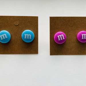 K&ouml;nnte beinhalten: Zwei Paar Ohrstecker. Ein Paar ist blau mit einem wei&szlig;en "M" auf jedem Ohrstecker. Das andere Paar ist lila mit einem wei&szlig;en "M" auf jedem Ohrstecker.