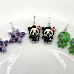 May include: Three pairs of dangle earrings featuring miniature figures: a pair of purple bats, a pair of black and white hooded figures with knives, and a pair of green stitched voodoo dolls, all attached to silver-toned metal earring hooks.