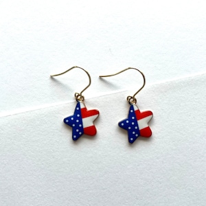 May include: A pair of gold dangle earrings with a star-shaped charm. The star is red, white, and blue with a pattern of white stars on a blue field.