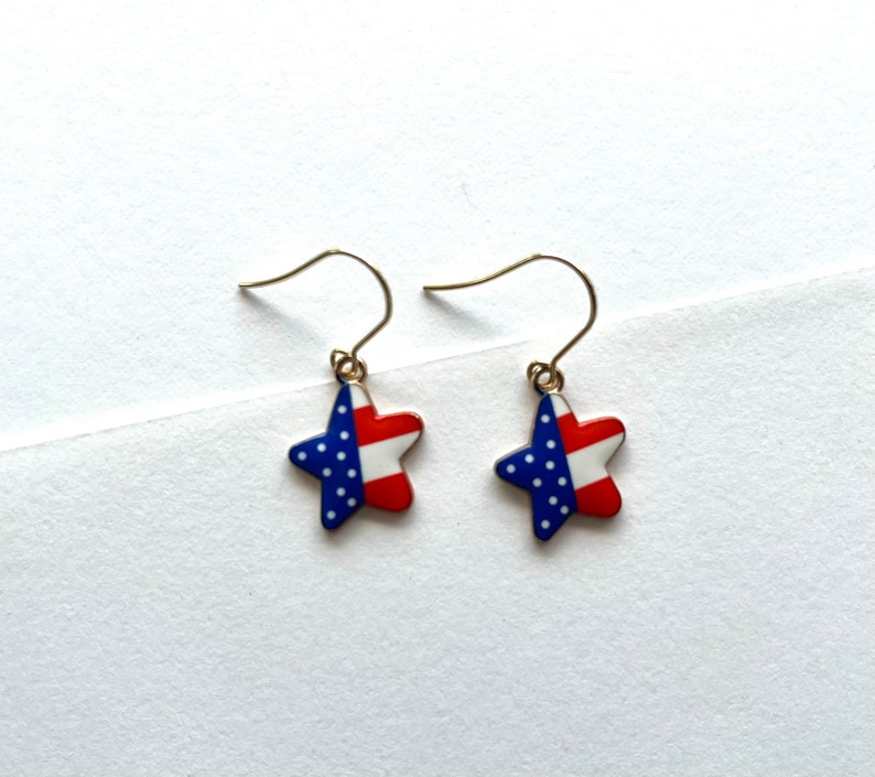 May include: A pair of gold dangle earrings with a star-shaped charm. The star is red, white, and blue with a pattern of white stars on a blue field.