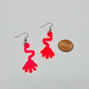 May include: A pair of neon pink dangle earrings featuring a hand shape with a wavy line leading up to it. The earrings are shown next to a US penny for scale.