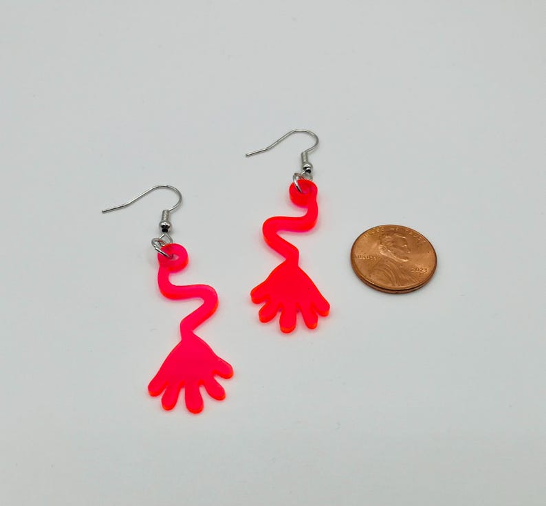May include: A pair of neon pink dangle earrings featuring a hand shape with a wavy line leading up to it. The earrings are shown next to a US penny for scale.