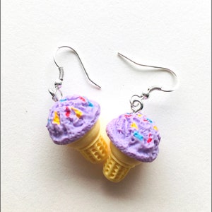 May include: A pair of dangle earrings featuring a purple ice cream cone with colourful sprinkles. The earrings are made of polymer clay and have silver hooks.