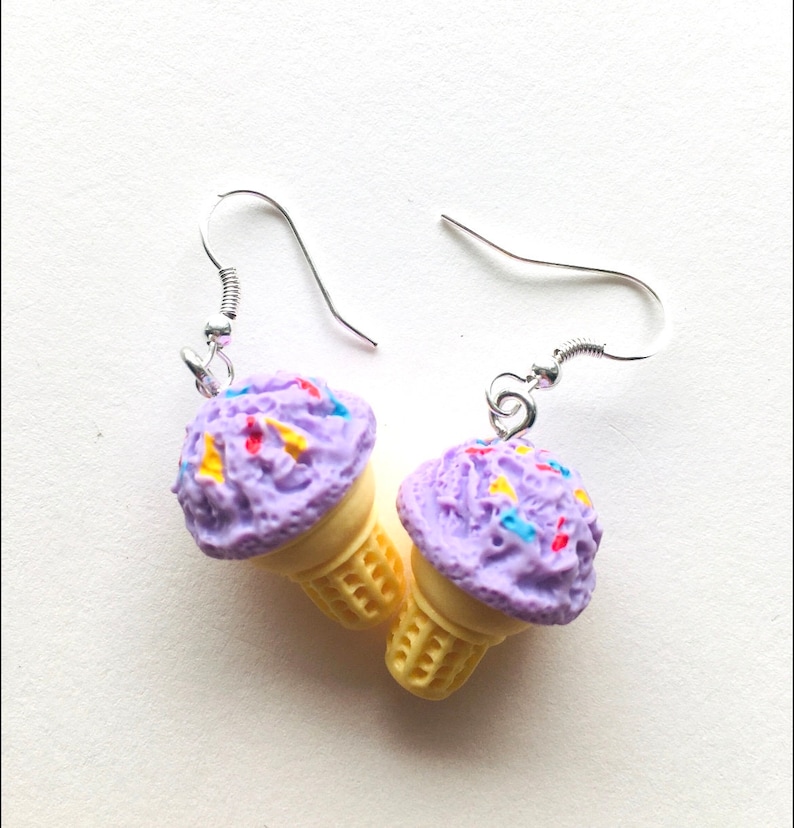 May include: A pair of dangle earrings featuring a purple ice cream cone with colourful sprinkles. The earrings are made of polymer clay and have silver hooks.