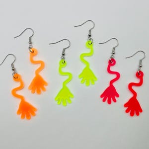 May include: A set of three pairs of neon-colored dangle earrings. The earrings are shaped like a hand with a wavy stem. The colors are orange, yellow, and pink.