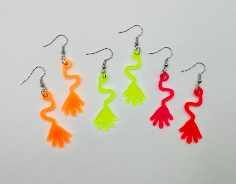 May include: A set of three pairs of neon-colored dangle earrings. The earrings are shaped like a hand with a wavy stem. The colors are orange, yellow, and pink.