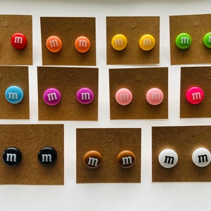 K&ouml;nnte beinhalten: Ein Set mit 12 Paar Ohrringen in verschiedenen Farben, darunter Rot, Orange, Gelb, Gr&uuml;n, Blau, Rosa, Lila, Braun, Schwarz und Wei&szlig;. Jeder Ohrring ist in Form eines M&M-Bonbons mit einem wei&szlig;en Buchstaben "M" auf der Vorderseite.