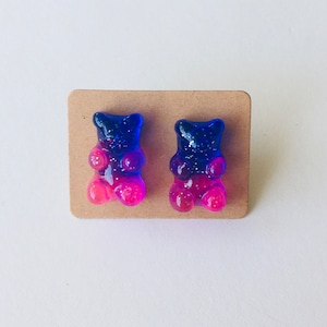 May include: A pair of gummy bear earrings with a blue to pink ombre effect. The earrings are made of resin and have a sparkly finish.