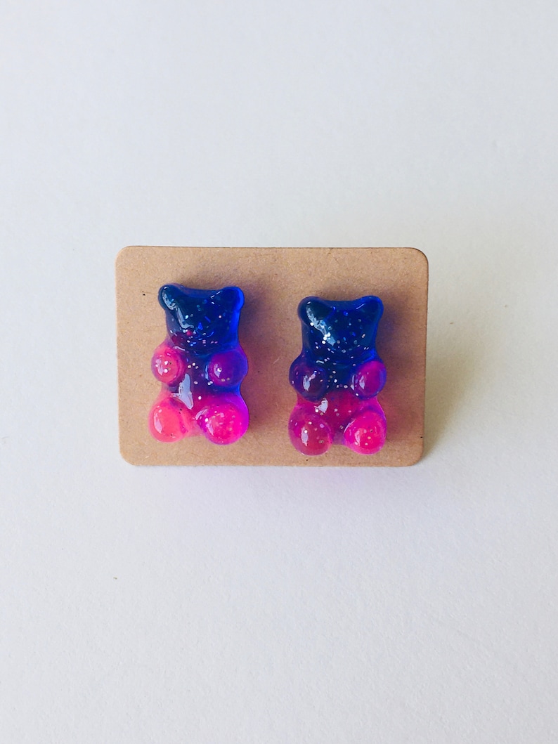 May include: A pair of gummy bear earrings with a blue to pink ombre effect. The earrings are made of resin and have a sparkly finish.