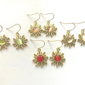 May include: A collection of gold-colored sun-shaped earrings with various colored centers, including green, pink, red, and cream. The earrings have a hook closure and are displayed on a white surface. The sun design features detailed rays.