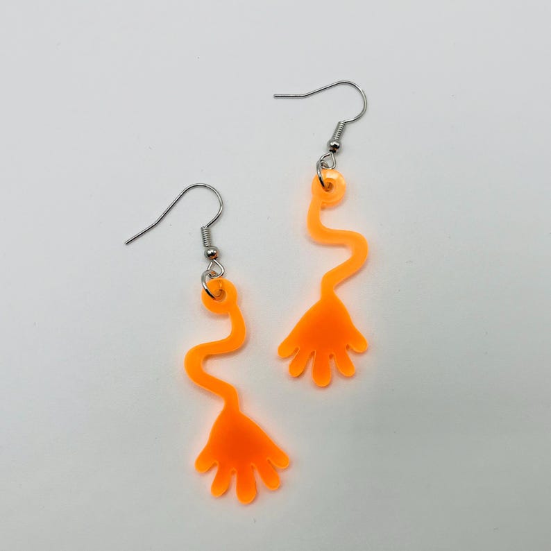 May include: A pair of orange dangle earrings with a hand shape at the bottom. The earrings are made of a translucent material and have a wavy design.