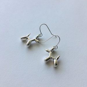 May include: A pair of silver balloon dog earrings. The earrings are made of metal and are shaped like balloon dogs. The earrings are hanging from silver hooks.