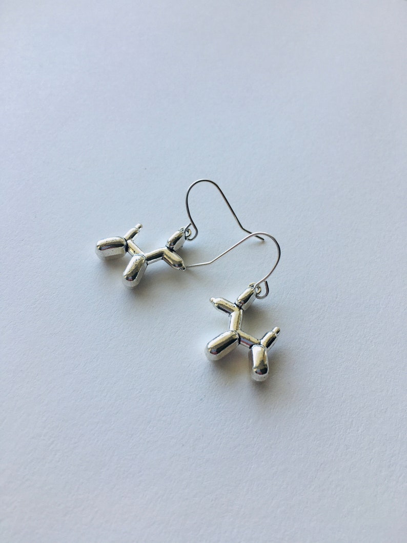 May include: A pair of silver balloon dog earrings. The earrings are made of metal and are shaped like balloon dogs. The earrings are hanging from silver hooks.