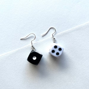 May include: A pair of dice earrings. One earring is black with a single white dot, the other is white with four black dots. The earrings have silver-colored fish hook ear wires.