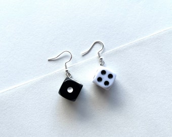 May include: A pair of dice earrings. One earring is black with a single white dot, the other is white with four black dots. The earrings have silver-colored fish hook ear wires.