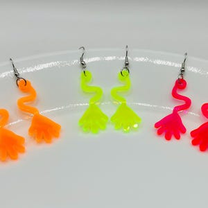 May include: Three pairs of neon colored dangle earrings. The earrings are shaped like a hand with a wavy line leading up to the hand. The earrings are available in pink, yellow, and orange.