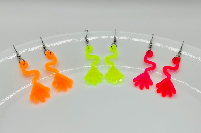 May include: Three pairs of neon colored dangle earrings. The earrings are shaped like a hand with a wavy line leading up to the hand. The earrings are available in pink, yellow, and orange.