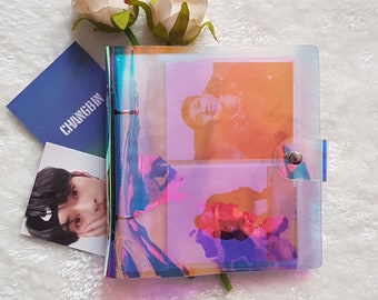May include: A clear iridescent binder with a silver ring mechanism. The binder has two clear plastic pockets that are filled with photos. The binder is sitting on a white fuzzy surface with two white flowers.