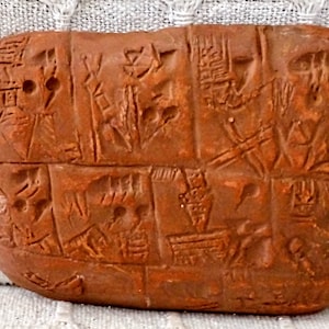 Puede incluir: Una tablilla de arcilla con escritura cuneiforme, una antigua forma de escritura utilizada en Mesopotamia. La tablilla es rectangular y tiene un color marrón rojizo.