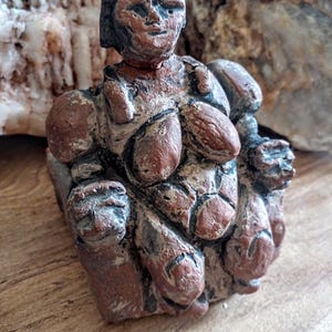 May include: A terracotta sculpture of a seated woman with a detailed, textured surface. The sculpture has a reddish-brown color with black accents.