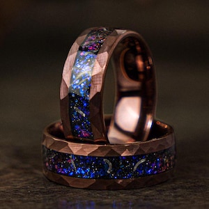 Op de afbeelding: Twee gestapelde ringen van roségoud wolfraamcarbide met een blauw en paars galaxy-patroon. De ringen hebben een facetgeslepen, diamantgeslepen ontwerp.
